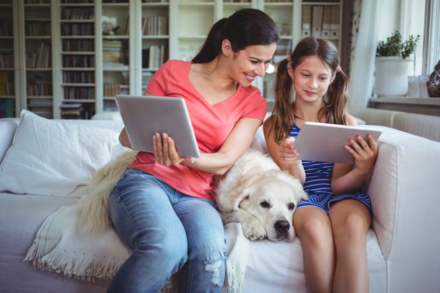Mother and daughter sitting on a couch in a cozy living room, both using digital tablets while their pet dog rests beside them. Perfect for illustrating family bonding, modern technology use, and home life. Ideal for advertisements, blog posts, or articles about parenting, technology in family life, or pet-friendly homes.