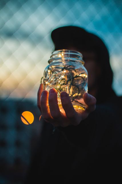 Person holding a jar filled with fairy lights during twilight in urban setting. Fairy lights glow softly inside the mason jar as surrounding background blurs with bokeh effect. Useful for concepts like dreams, imagination, creativity, modern lifestyle, or urban aesthetics.