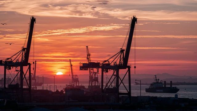 Gantry cranes silhouetting against vibrant harbor sunset with cargo ship on horizon