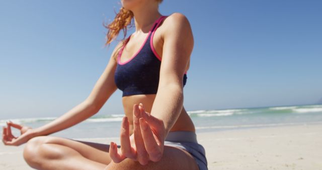 Young woman sitting cross-legged on a sandy beach practicing yoga and meditation in bright, sunny weather. Ideal for use in wellness, fitness, yoga retreats, beach vacation promotions, mindfulness and relaxation campaigns.
