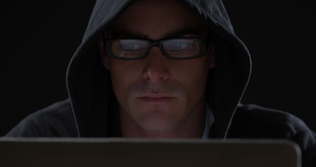 This image of a person wearing a hoodie and glasses using a laptop in a dark room can be used for cybersecurity, hacking narratives, anonymous online activities, and discussions on internet privacy. It is ideally suited for illustrating articles, blogs, or advertisements related to cybercrime, data protection, and online security.