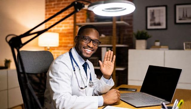 Smiling doctor waving during telemedicine consultation at home office desk with laptop