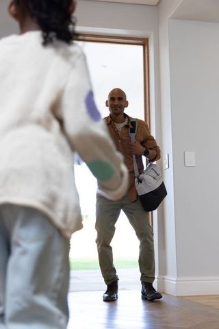 Diverse father returning home greeting excited daughter at doorway carrying duffel bag