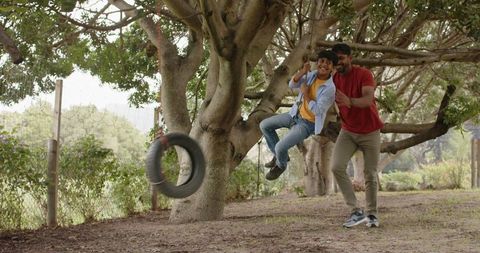 Father pushing son on tire swing in shaded garden