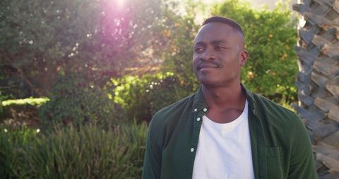 African american man standing in sunlit garden with relaxed smile and casual green shirt