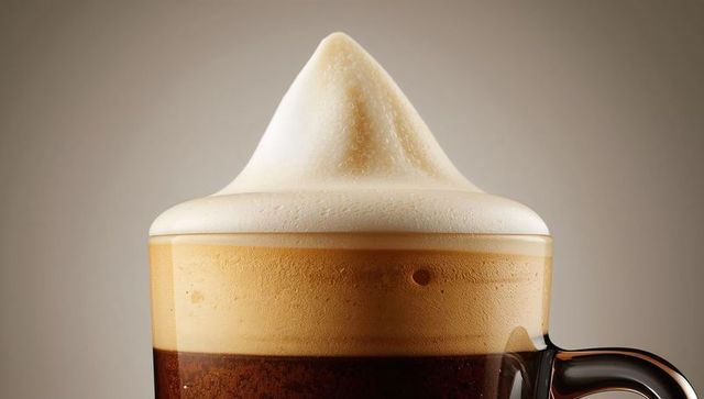 Frothy latte macchiato in clear glass mug with creamy peak and condensation, studio-lit close-up