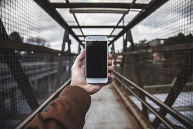 Person holding smartphone with blank screen on metal bridge. Suitable for technology, mobile app promotion, and travel-related content.