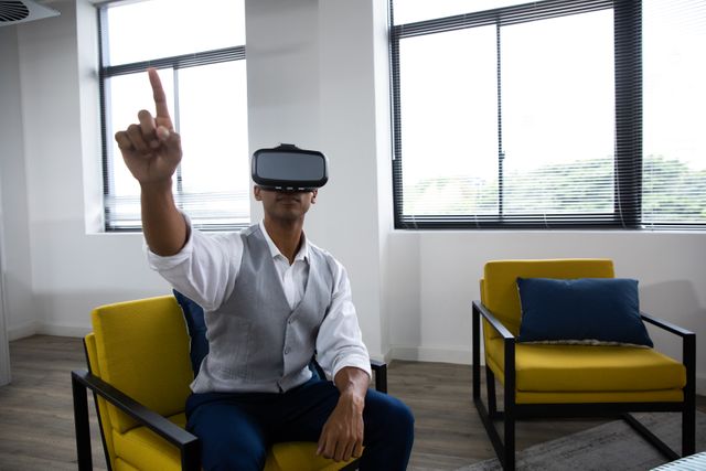 Front view of a young African American man sitting wearing a VR headset and with one hand raised and finger pointing in the lounge area of a creative business.