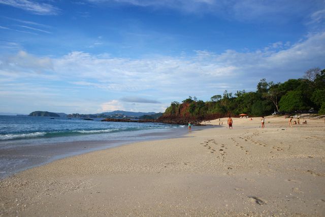 Calm sandy beach with clear blue sky and lush greenery in the background, offers a serene environment for relaxation and outdoor activities. Ideal for travel agencies, beach resort advertisements, vacation planners, and nature-inspired designs.
