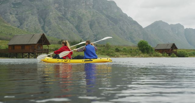 Couple kayaking on serene mountain lake, wearing outdoor attire. Water reflects scenic mountain and cabins in background under cloudy sky. Ideal for promoting outdoor activities, adventure tours, travel destinations.