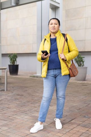 Mature woman standing in yellow puffer jacket holding smartphone in modern urban plaza