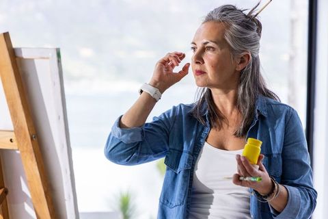 Mature woman painting at easel holding yellow paint bottle contemplating waterside studio