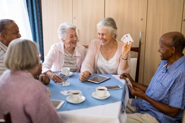 Group of senior people sitting around a table, playing cards and enjoying tea. They are smiling and engaging in conversation, creating a warm and friendly atmosphere. Ideal for use in content related to elderly care, retirement communities, social activities for seniors, and promoting a positive and active lifestyle for older adults.