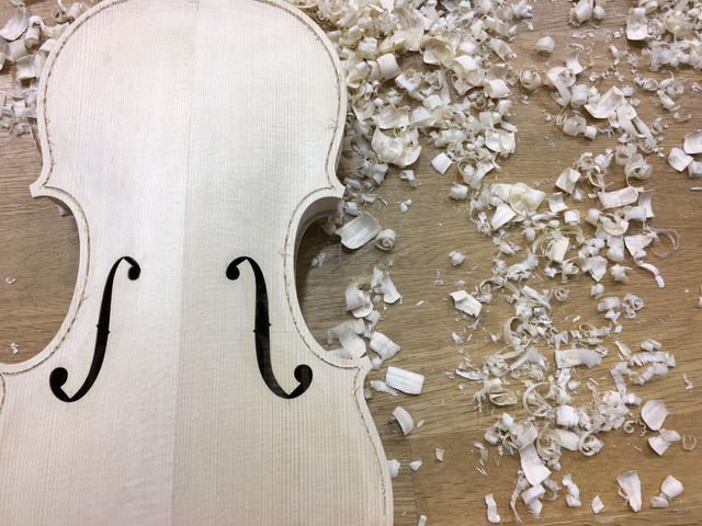 Partially crafted violin lying on a wooden workbench surrounded by wood shavings. Perfect for depicting handmade musical instrument creation, craftsmanship, and woodworking art. Useful for articles about traditional violin making, artisan skills, and music craftsmanship.