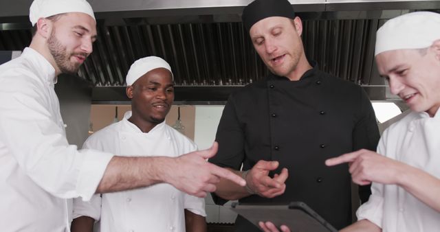 Four chefs are gathered in a professional kitchen, collaborating and smiling while they look at a tablet. The kitchen staff is dressed in white uniforms and chef hats, highlighting a teamwork environment. This image is ideal for articles and materials focusing on teamwork, culinary education, modern technology in the kitchen, or professional cooking environments.