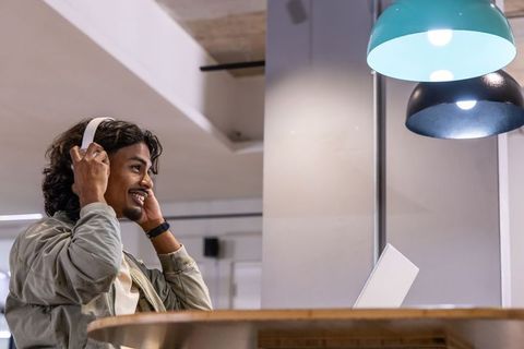 Young south asian man adjusting headphones at coworking high table with laptop, teal lamps