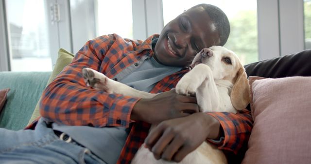 Happy african american man sitting on couch ad playing with pet dog at home. Lifestyle, pets and domestic life, unaltered.