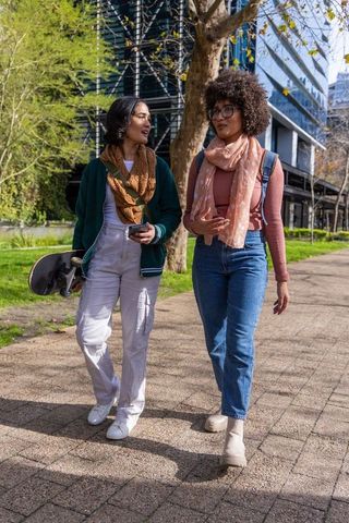 Asian and african american women walking in urban park carrying skateboard and backpack