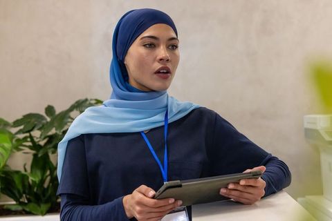 Female receptionist wearing blue hijab standing behind modern front desk holding tablet