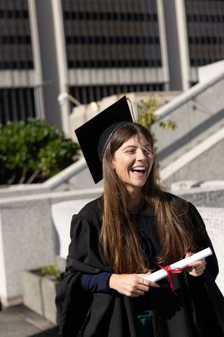 Joyful graduate laughing in mortarboard and gown holding diploma on sunlit campus plaza