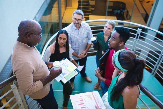 Mature businessman explaining graphical data to a multiracial team of coworkers on a staircase in an office. Ideal for depicting teamwork, corporate meetings, business discussions, and professional collaboration in a diverse workplace setting.