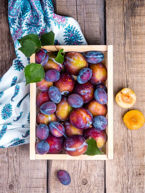 Fresh colorful plums are displayed in a wooden crate on a rustic wooden table, surrounded by plums and a patterned cloth. This image is perfect for blogs or articles about organic farming, healthy eating, autumn harvest, local markets, or culinary recipes. It can also be used for promotional material in food-related businesses such as marketplaces, grocery stores, and restaurants.