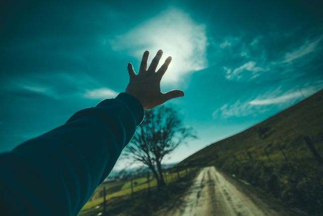 Depicts a hand reaching towards the bright sky on a rural dirt road, symbolizing freedom, aspiration, and exploration. Suitable for use in promotional materials, inspirational content, websites, blogs about nature, travel brochures, or adventure-themed projects.