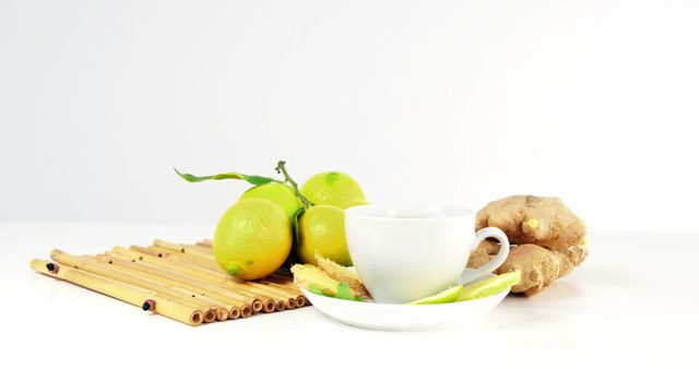 Image depicting fresh lemons and ginger root with a white tea cup on a bamboo mat, set against a white background. Ideal for use in health and wellness articles, food and drink blogs, natural remedy guides, and cooking magazines. Perfect for promoting a healthy lifestyle and detox tea benefits.