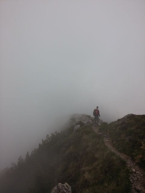 Man walking on a foggy mountain ridge, suggesting themes of adventure and solitude. Suitable for use in travel and adventure blogs, advertisements for outdoor gear, and inspirational content highlighting exploration and nature. The image can evoke a sense of mystery, challenge, and serenity in the great outdoors.