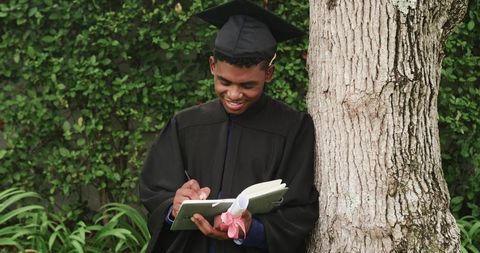 African American Graduate Writing Notebook While Leaning on Tree and Holding Diploma