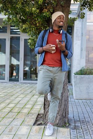 African american man leaning on tree holding coffee and smartphone urban beanie headphones