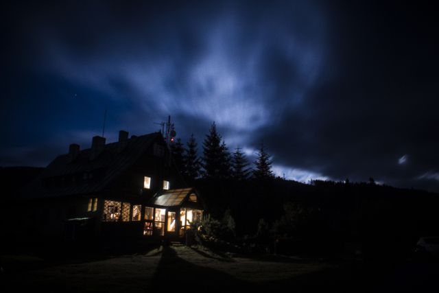 Cozy mountain cabin with illuminated windows under a dark, cloudy night sky. Highlights the tranquility and solitude of remote living, appealing to those seeking adventure or looking for serene escape imagery. Perfect for use in outdoor recreation promotions, travel brochures, and lifestyle magazines aimed at nature enthusiasts.