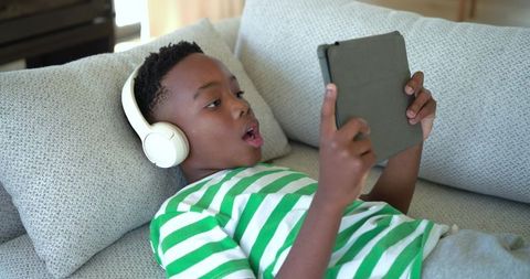 School-age african american boy reclining on couch wearing headphones using tablet for learning