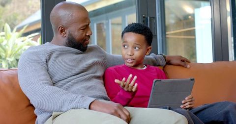 African american father and son discussing schoolwork on tablet while relaxing on couch