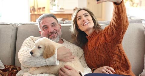 Smiling couple taking cozy selfie on sofa with golden retriever puppy in knit sweaters