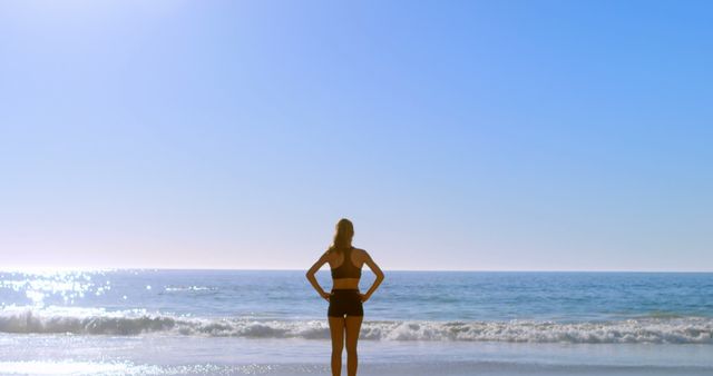 Rear view of young woman standing on sandy beach, looking at ocean. Ideal for travel and leisure blogs, vacation advertisements, wellness and relaxation content, and outdoor activity promotions.