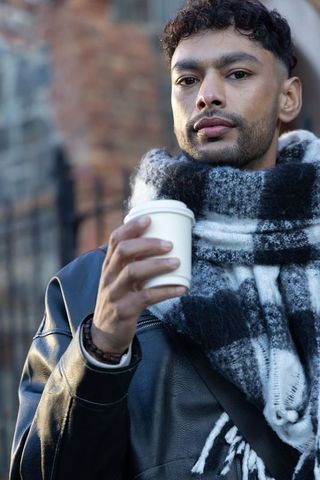 Young man wearing leather jacket and checkered scarf holding takeaway coffee cup