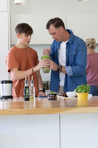 Father teaching teenage son making green smoothie on wooden island in modern kitchen