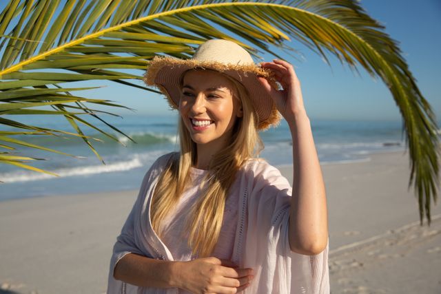 Caucasian woman enjoying time at the beach on a sunny day, wearing sun hat with palm tree sea in the background. Holiday relaxing summer wellbeing.