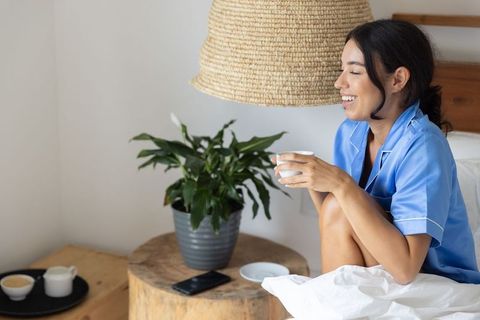 Woman enjoying morning tea in blue pajamas on cozy bed under woven lampshade smiling softly