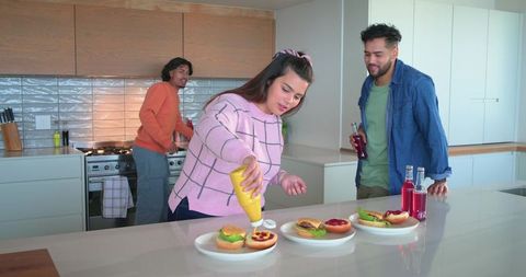 Multicultural friends preparing burgers at kitchen island, squeezing mustard, casual drinks