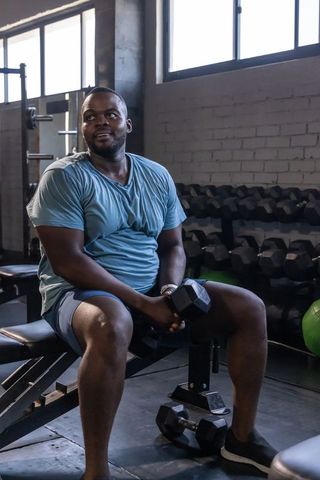 African american man sitting on bench holding dumbbell in gym practicing strength training