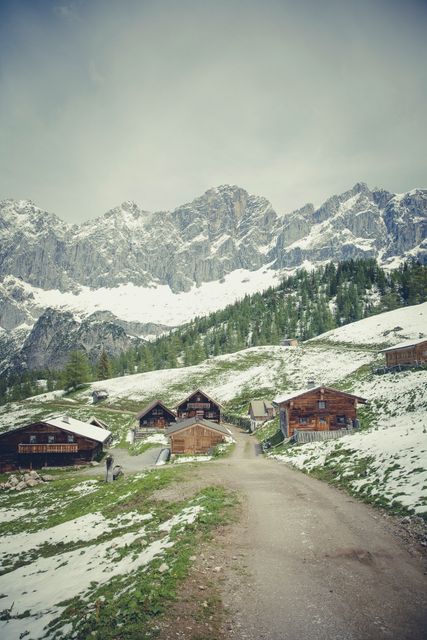 Scenic mountain village with charming wooden cabins nestled among snow-capped peaks and a winding trail. Ideal for content related to nature, travel destinations, adventure tourism, rural tranquility, and outdoor activities.