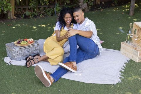 Couple relaxing on picnic blanket sharing smartphone and fruit platter in backyard
