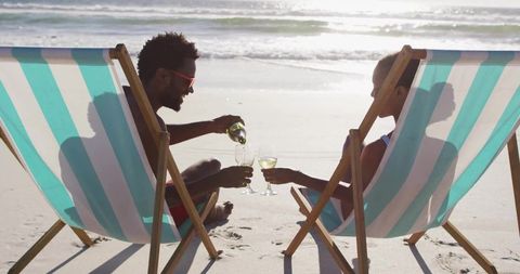 Couple toasting white wine in striped deck chairs on sunlit beach shoreline