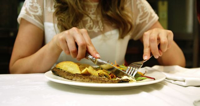 Woman using knife and fork while enjoying a gourmet meal in a formal restaurant. Ideal for use in articles or advertisements about dining etiquette, food reviews, lifestyle blogs, or restaurant promotions.