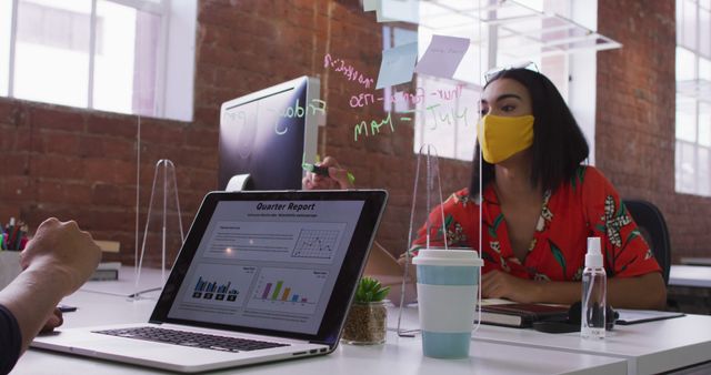 Biracial businesswoman writing on sneez screen sitting in front of computer wearing face mask. independent creative design business during covid 19 coronavirus pandemic.