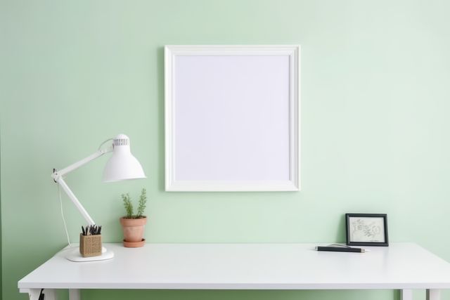 A clean, minimalistic desk set up in a home office featuring a blank frame on a pastel green wall, a desk lamp, small plants in terracotta pots, a pen, and a couple of small desk accessories. Ideal for articles and blogs on interior design, workspace organization, minimalism, and modern home decor.