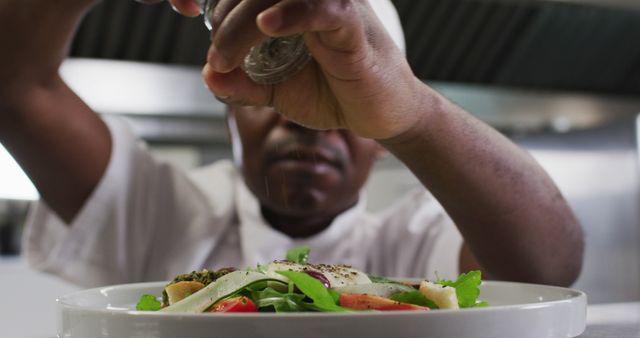 Close-up of chef adding salt to fresh salad in a modern kitchen. Ideal for articles, blogs, and advertisements related to food preparation, culinary skills, healthy eating, and professional kitchen environments.
