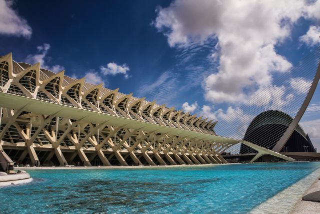 This image captures the modern and futuristic architecture of the City of Arts and Sciences in Valencia, Spain. The buildings reflect on the blue water beneath a sky dotted with clouds. This can be used to promote travel and tourism, for architectural design inspiration, or as a representation of innovation and modern urban landscapes.